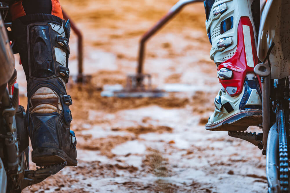 Two Motocross riders in protective gear with boots on a muddy track, ready to race