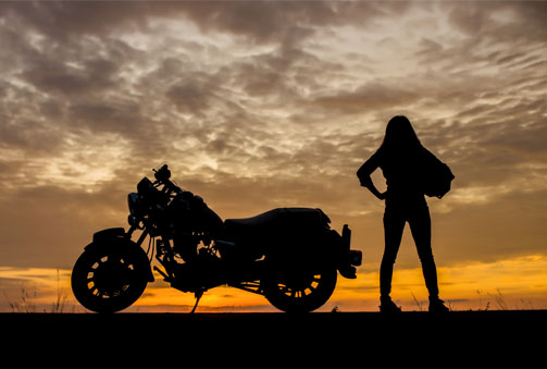 The silhouette of a woman standing next to a motorcycle against the sunset