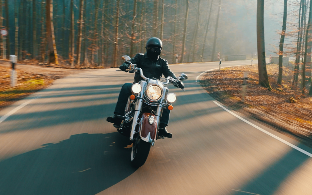 Rider on classic motorcycle cruising on forest road, autumn leaves on ground.