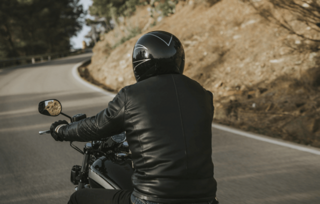A motorcyclist viewed from behind, wearing a black helmet and leather jacket while riding on a winding road