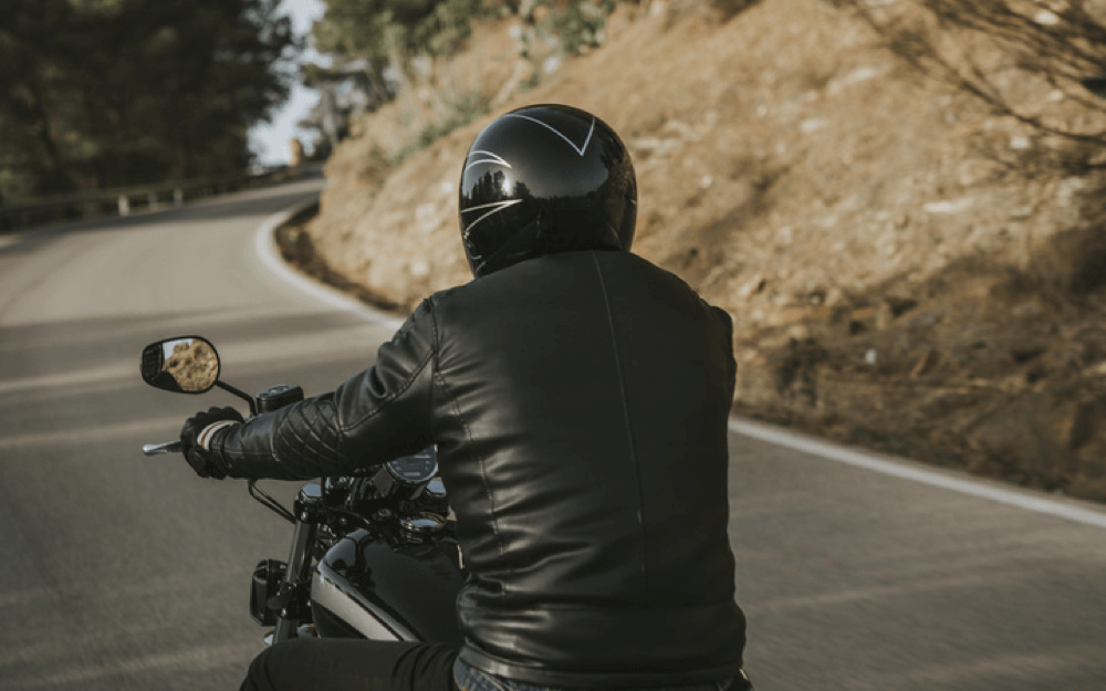 A motorcyclist viewed from behind, wearing a black helmet and leather jacket while riding on a winding road