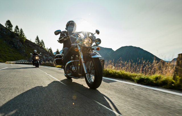 Two motorcyclists with touring bikes riding on a mountain road under a clear sky with the sun shining brightly.