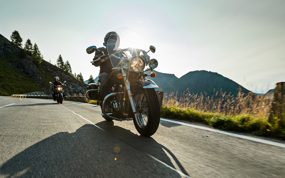 Two motorcyclists with touring bikes riding on a mountain road under a clear sky with the sun shining brightly.