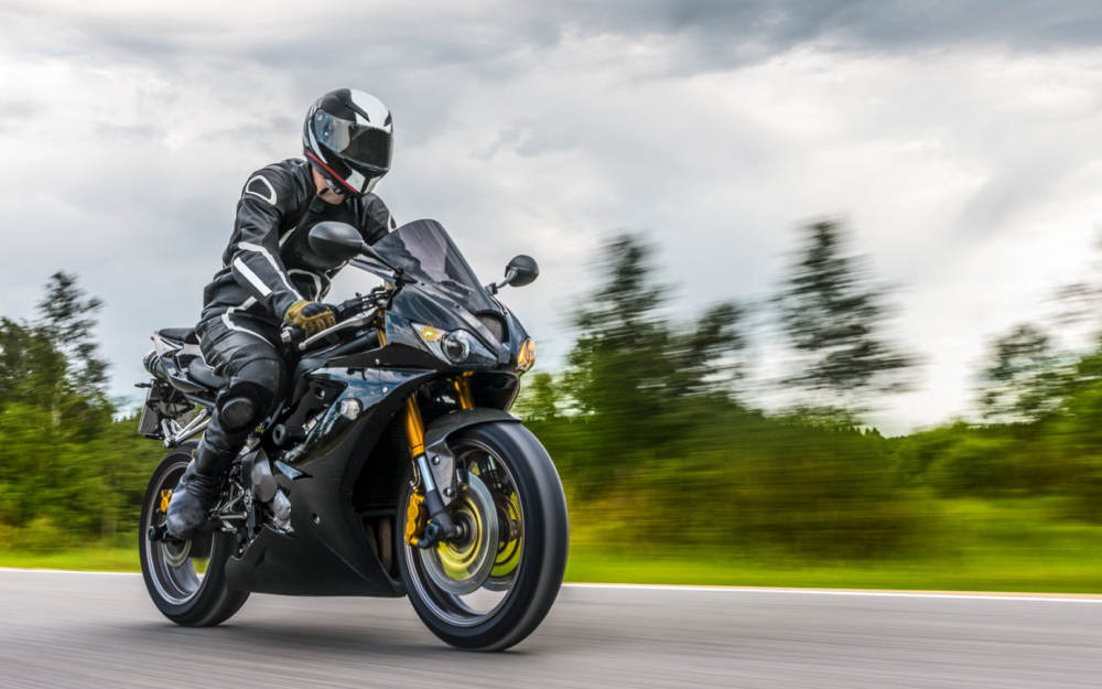 A motorcyclist in full black leather racing suit and helmet on a sport bike, with a blurred background indicating rapid movement.