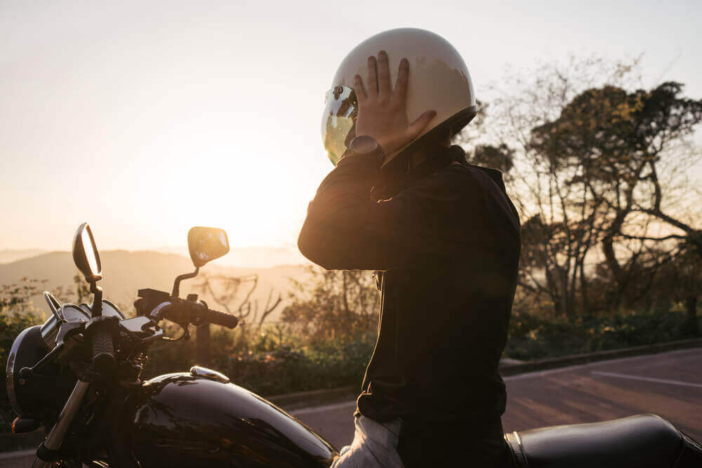 A motorcyclist in a white helmet taking off his helmet while sitting on a motorcycle