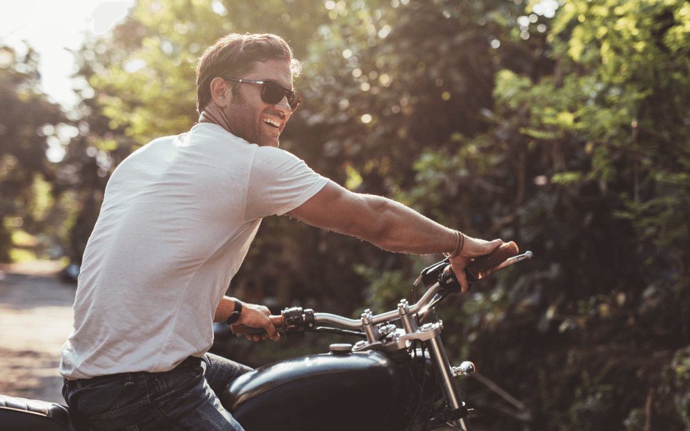 A cheerful man in sunglasses riding a classic motorcycle, with sunlight filtering through the green foliage