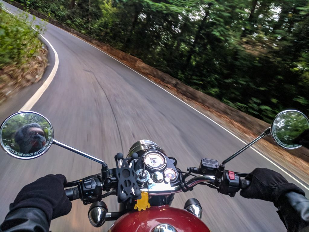 First-person view of a motorcyclist riding on a winding road through a forest, reflection visible in side mirrors.