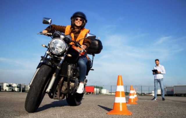 A smiling woman in a helmet and orange safety vest riding a motorcycle with an instructor and traffic cones in the background