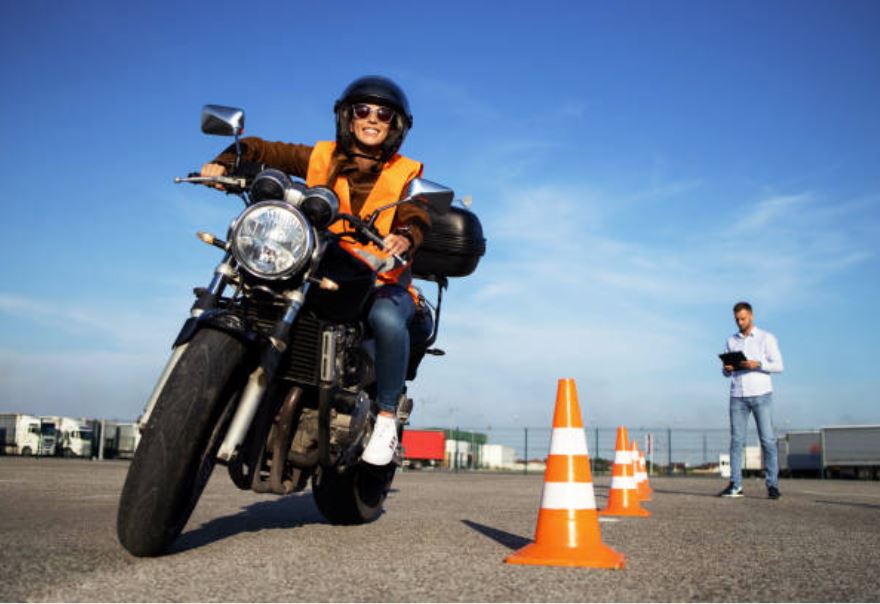 A smiling woman in a helmet and orange safety vest riding a motorcycle with an instructor and traffic cones in the background